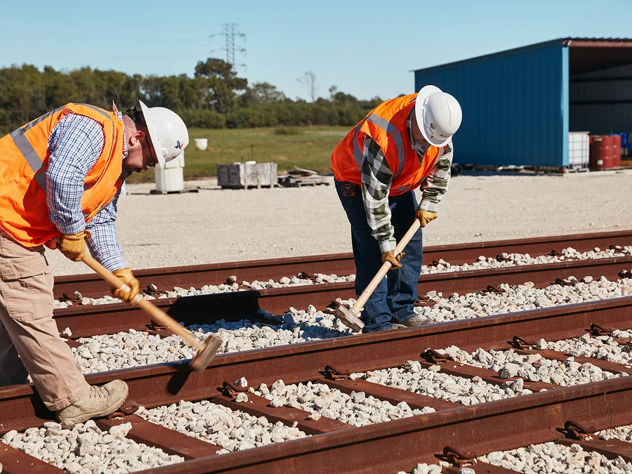 Two workers in safety gear use sledgehammers to work on railroad tracks outdoors, surrounded by gravel and construction materials.
