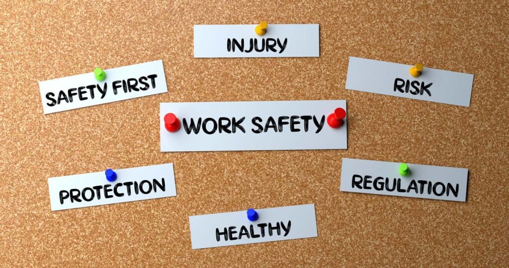 Bulletin board with notes reading "WORK SAFETY," surrounded by other notes: "SAFETY FIRST," "INJURY," "RISK," "PROTECTION," "REGULATION," and "HEALTHY," pinned with colored tacks.