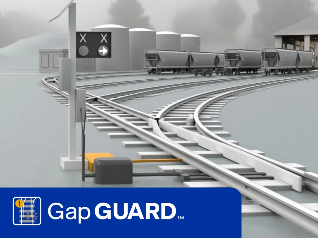 A railway track with a signal, a device labeled "GapGUARD," and freight cars in the background under cloudy skies.