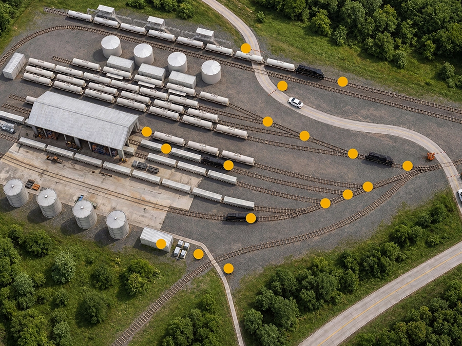 Aerial view of a rail yard with multiple train tracks, stationary tanker cars, fuel storage tanks, and a small building, bordered by greenery and roads.