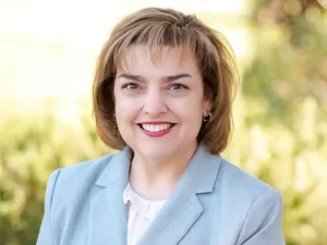A woman with short light brown hair wearing a light blue blazer and white top smiles outdoors in front of greenery.
