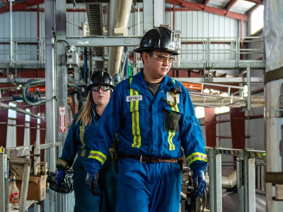Two industrial workers in blue protective uniforms and hard hats walk inside a large facility with metal pipes and equipment visible in the background.
